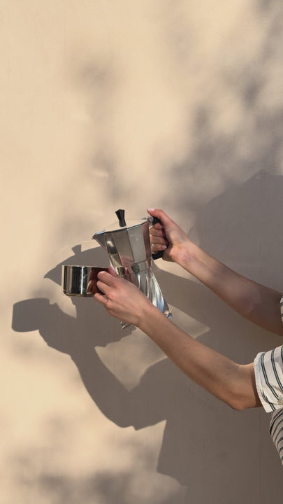 A hand pours espresso from the Turbo Moka into a small glass cup against a sunlit wall