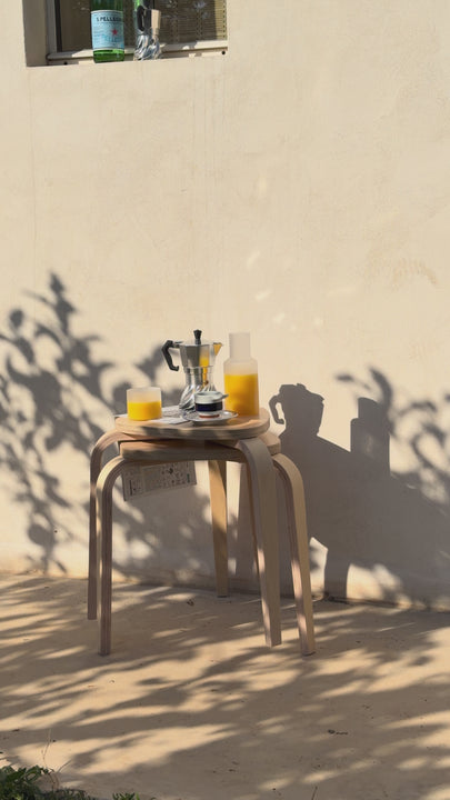Turbo Moka and a carafe of orange juice on a wooden stool against a white wall, dappled leaf shadows moving across the scene