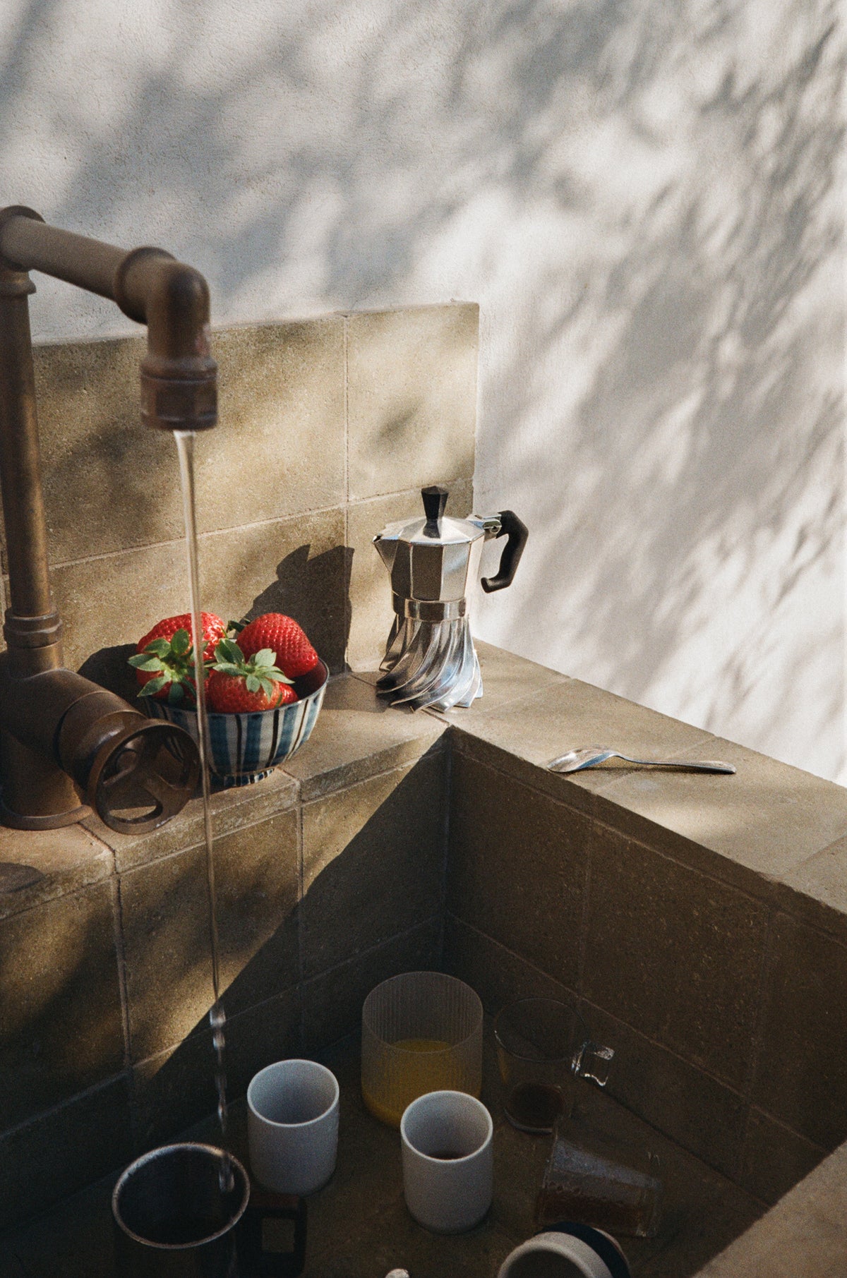 Turbo Moka beside a bowl of strawberries at an outdoor stone sink, water running from a brass tap into cups below